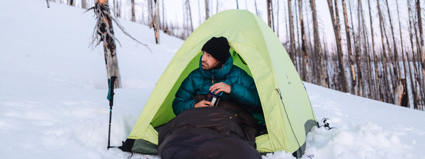 Man winter camping in snowy forest, staying warm inside a lightweight tent with a Zenbivy Light Bed sleeping system and insulated jacket.