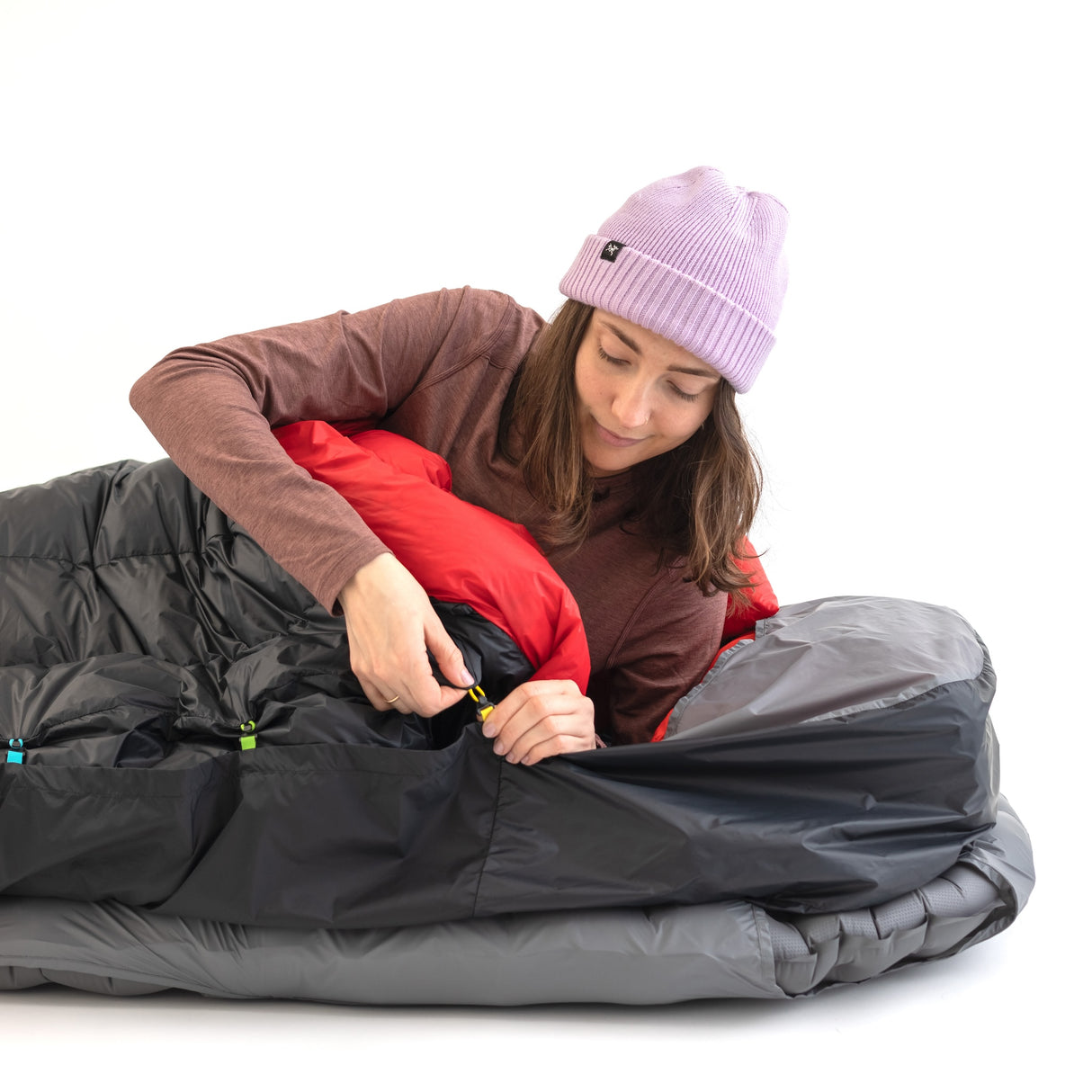 Woman securing a Zenbivy sleeping bag using the color-coded clip system, lying on a gray mattress with a red and black quilt.