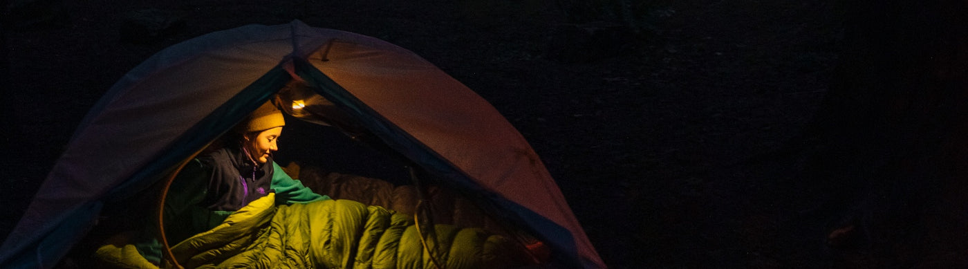 A person illuminated by a warm light sits inside a tent at night, cozily tucked into a green sleeping quilt. The surrounding forest is dark, creating a peaceful camping scene.