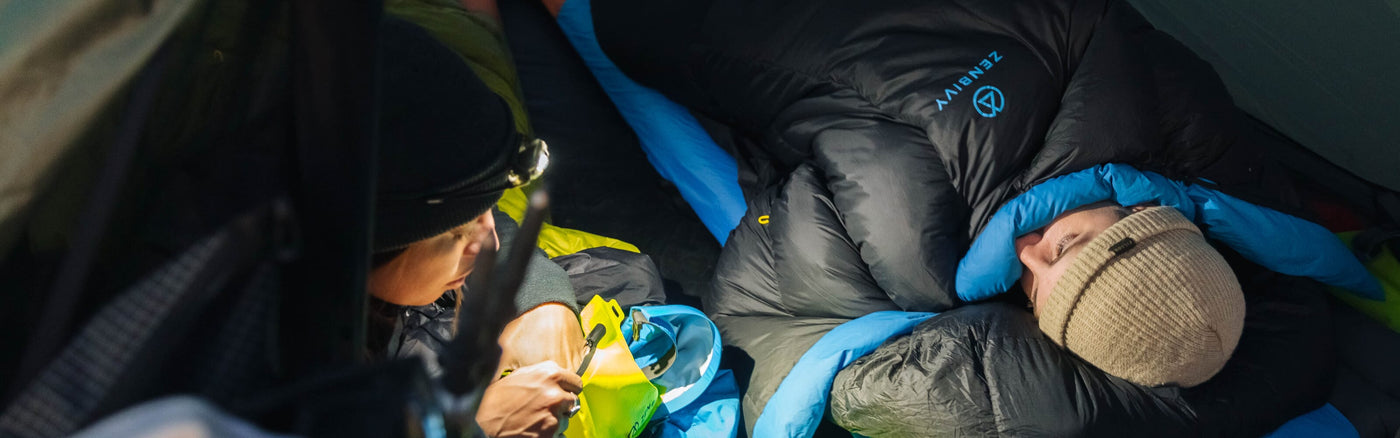 Two campers inside a tent at night, one person wearing a headlamp organizing gear, while the other is bundled in a Zenbivy sleeping bag, resting.