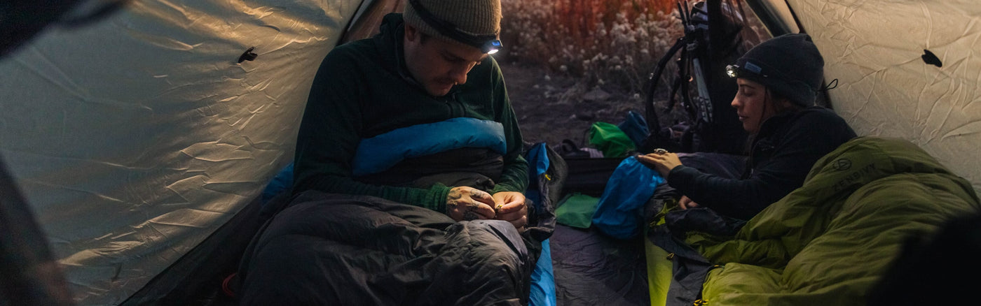 Two campers inside a tent at dusk, organizing gear and adjusting a Zenbivy double sleeping system. Both wear headlamps for light.