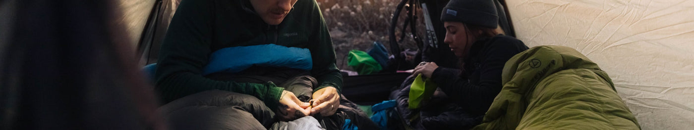 Two campers inside a tent, surrounded by sleeping bags and gear, preparing for the night. One person is adjusting their Zenbivy sleep system, while the other organizes equipment.