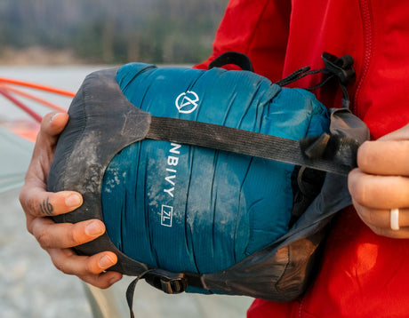 Close-up of hands adjusting the strap on a blue Zenbivy 7L compression sack, with a person wearing an orange jacket visible in the background.