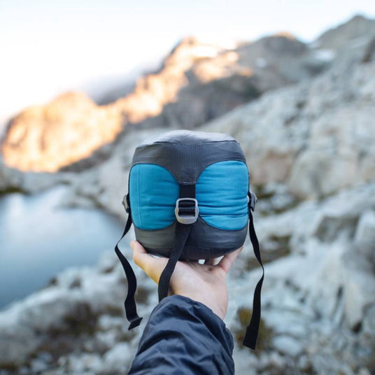 Person holding a compact, blue and black Zenbivy stuff sack with compression straps in front of a mountain lake backdrop.