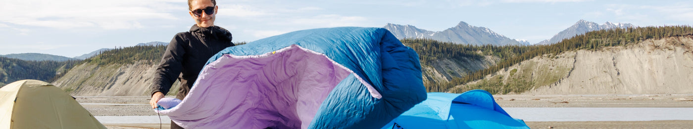 Person smiling while shaking out a Zenbivy Core Quilt with a purple lining at a campsite, surrounded by tents, mountains, and a river in the background.