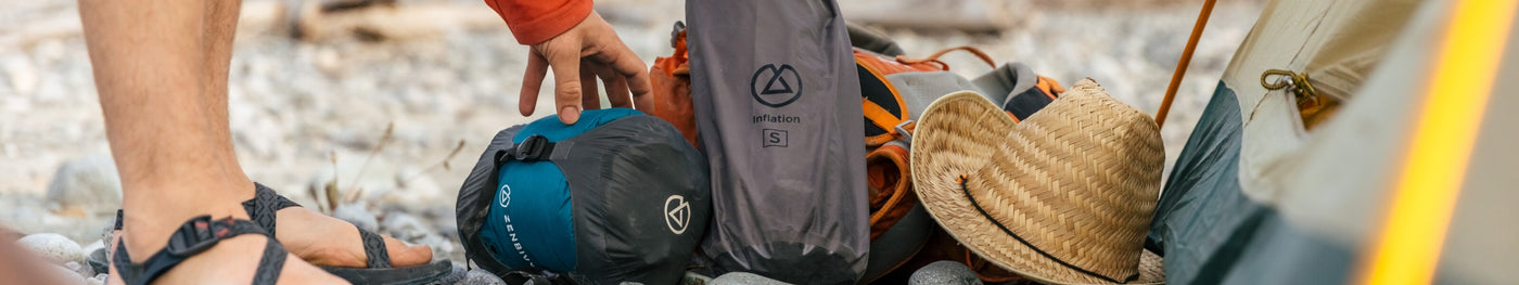 Close-up of a camper’s hand packing a Zenbivy compression sack next to an inflation bag, backpack, and straw hat at a campsite with rocky ground.