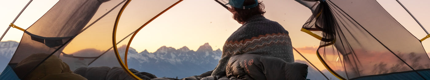 Camper sitting inside a mesh tent at sunrise, wrapped in a blanket and surrounded by Zenbivy sleep gear, overlooking a mountain range under a colorful sky.