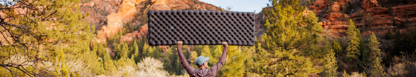 Person standing outdoors in front of red rock formations and pine trees, holding a Zenbivy inflatable sleeping pad overhead.