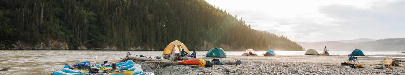 Panoramic view of a riverside campsite at sunset with multiple tents set up along a rocky shore. Several colorful inflatable rafts and kayaks are pulled up near the water, while camping gear and chairs are scattered across the beach. A dense forest and distant mountains rise in the background under a partly cloudy sky.
