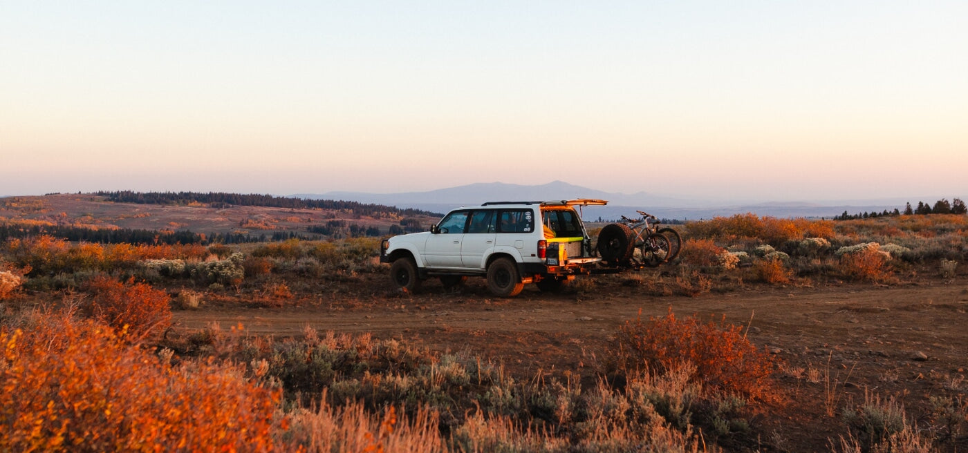 White SUV parked on a dirt road with bikes mounted on the back, overlooking a scenic, autumn-colored landscape at sunset.