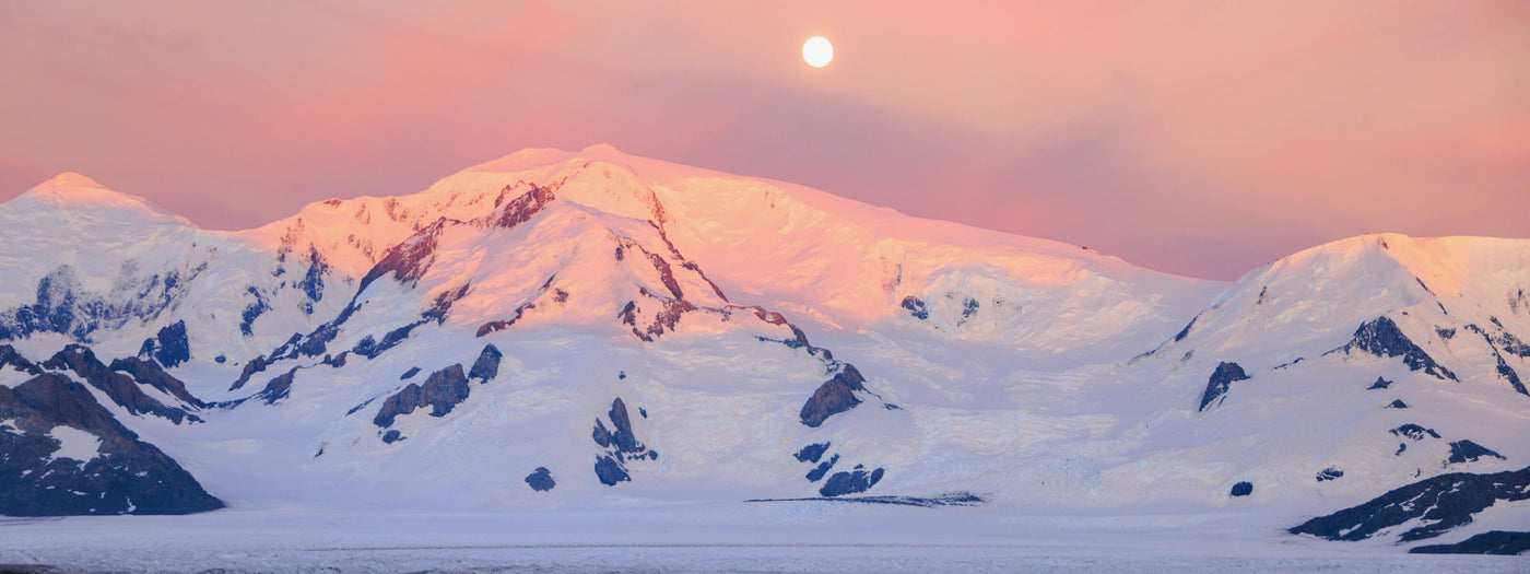 Snow-covered mountain range glowing under a pink sunrise sky with the moon overhead, perfect backdrop for winter camping and alpine adventures.