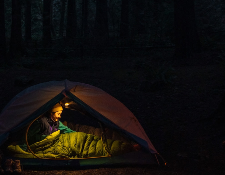 Girl in her sleeping bag inside her tent. It's dark out and she is illuminated by a headlamp. 