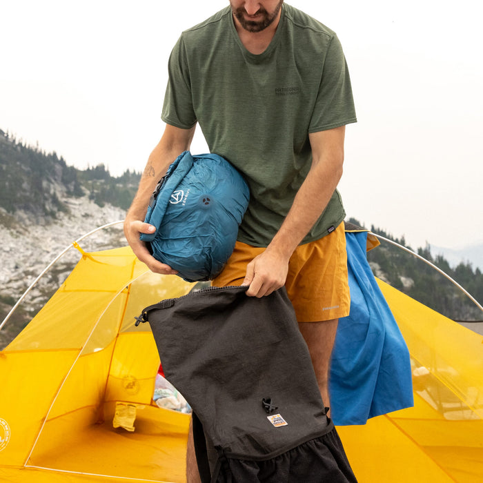 Man holding a blue Zenbivy Pack Liner, stuffing it into his backpacking outdoors with a mountainous background and yellow tent behind him