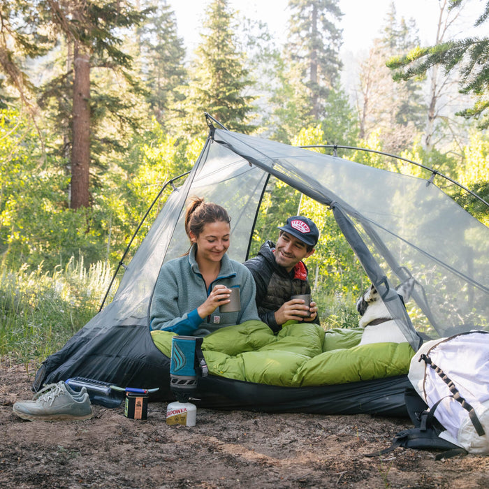 Couple with their dog sitting in a Zenbivy Double bed inside a grey tent drinking coffee in the morning, surrounded by trees
