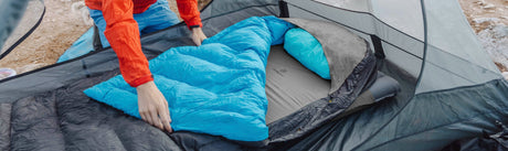 Person setting up a blue sleeping bag inside a tent outdoors.