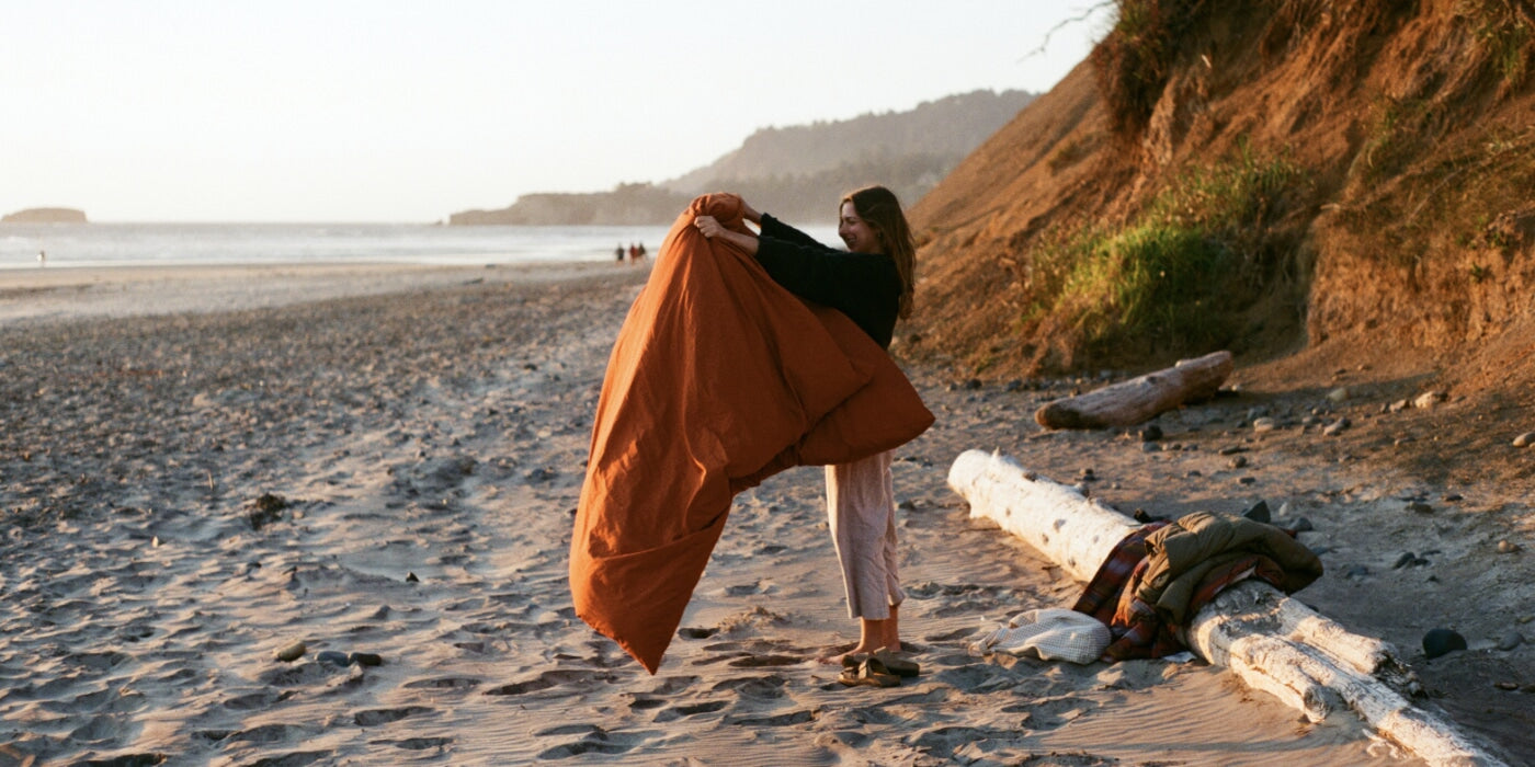 A person smiling while shaking out a Zenbivy Duvet Cover on a sandy beach at sunset, with cliffs and driftwood in the background.