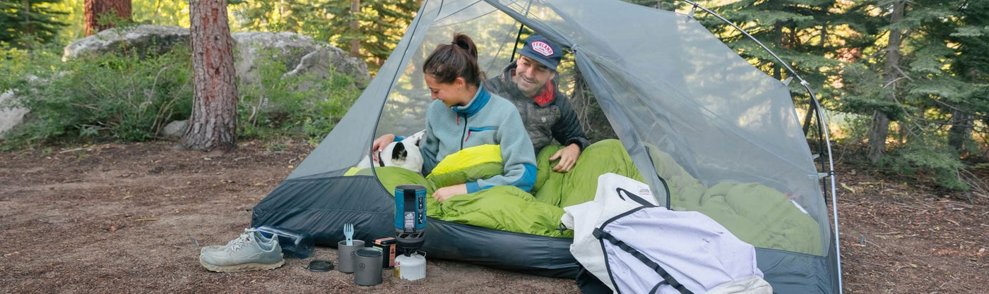Two people camping outdoors, sitting inside a tent with camping gear around them.