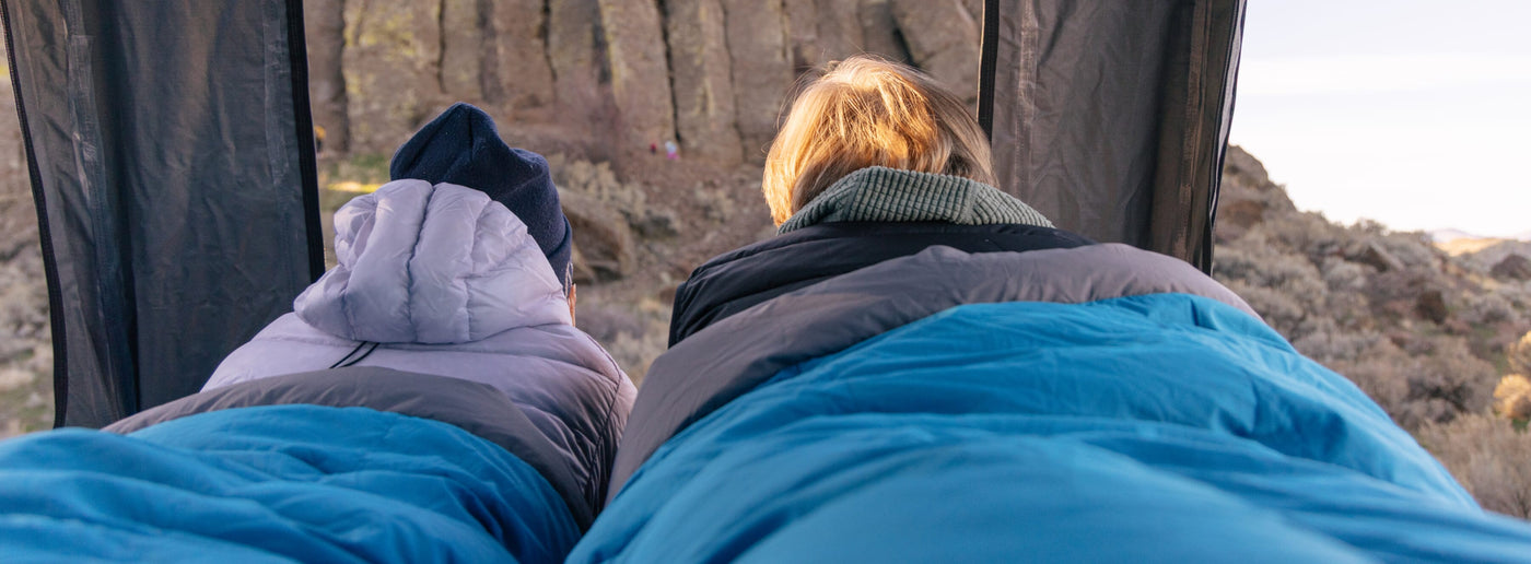 Two people lying in a blue Zenbivy Double Quilt inside a rooftop tent, looking out over a rocky landscape
